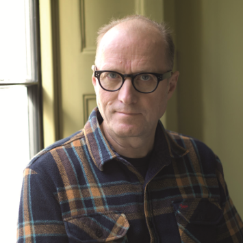 Ade Edmondson in checked shirt standing in front of window