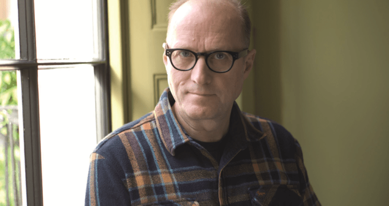 Ade Edmondson in checked shirt standing in front of window