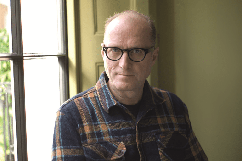 Ade Edmondson in checked shirt standing in front of window