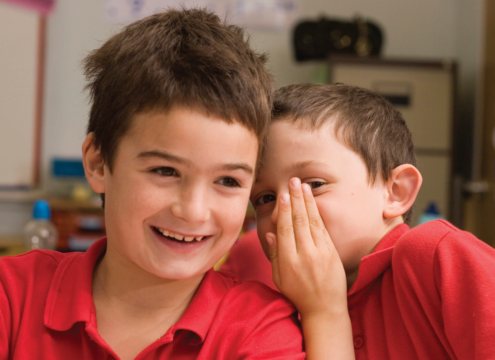 Two young boys in red school uniform tops whispering to each other