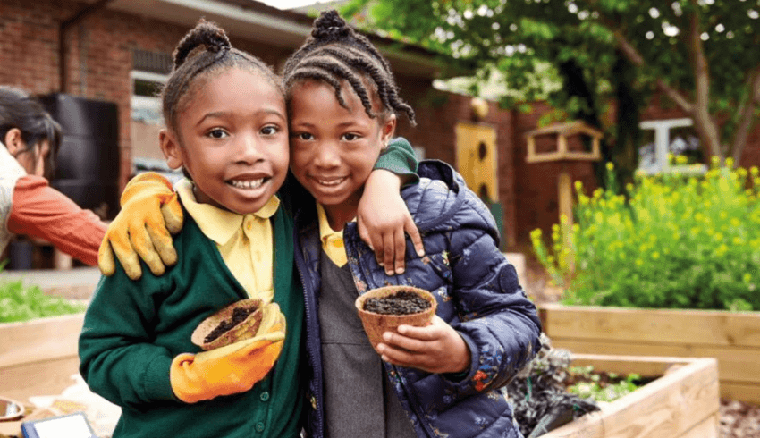 Two young pupils with arms around each other, holding plant pots in a garden