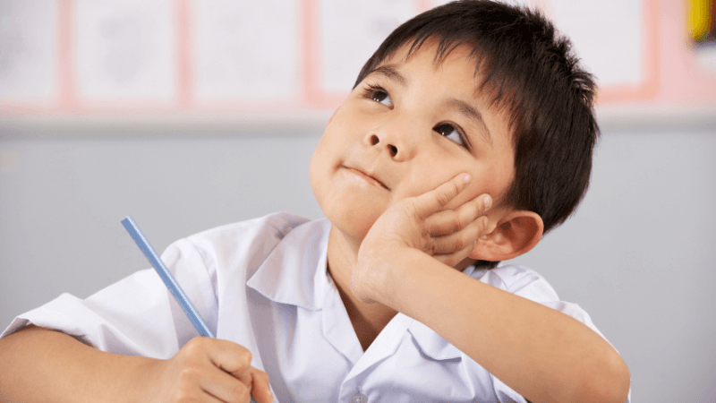Young boy with black hair and white shirt looking upwards and holding a pencil