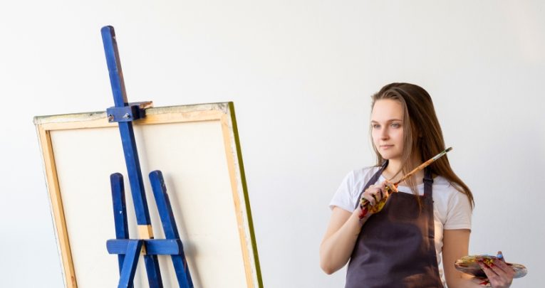 A female student pictured holding a paintbrush in front of an eisel
