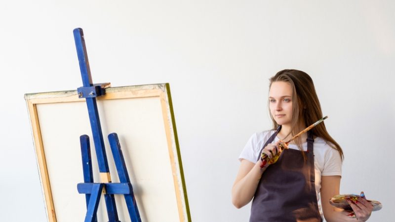 A female student pictured holding a paintbrush in front of an eisel