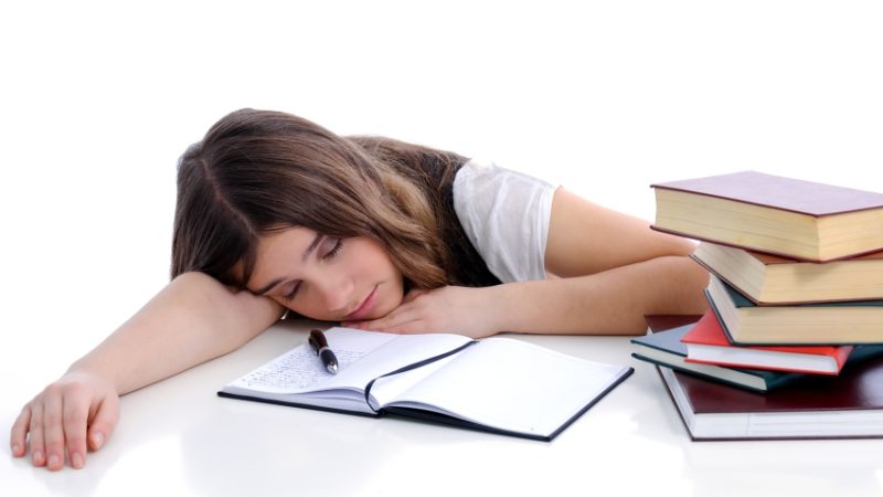 Photo of a lethargic and tired-looking female student surrounded by textbooks