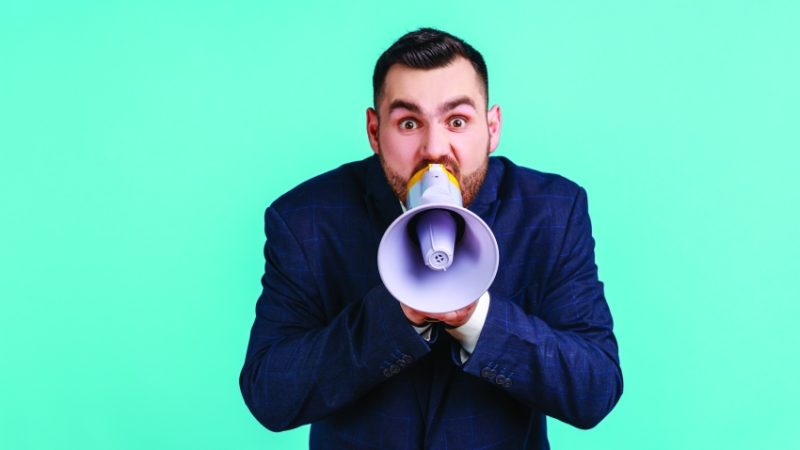 Photograph of a besuited man talking into a loudhailer
