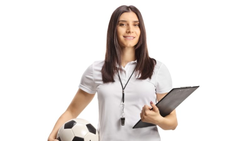 photo of woman in sportswear holding a football in one hand and a clipboard in the other