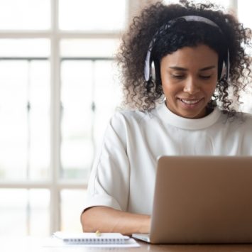 photo of young female teacher sat at a desk with a laptop while wearing a pair of headphones