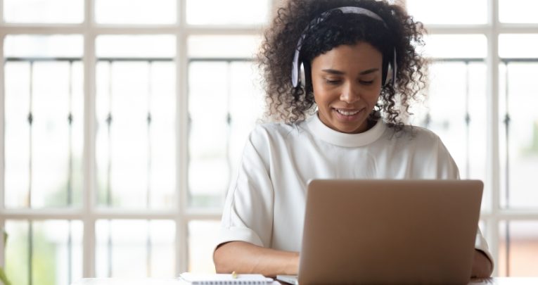 photo of young female teacher sat at a desk with a laptop while wearing a pair of headphones