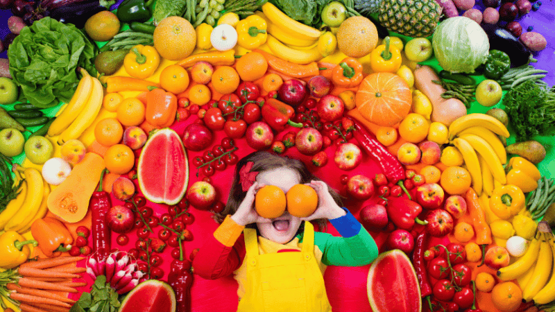 Kid surrounded by rainbow of fruit for Healthy Eating Week