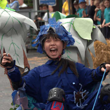 Girl in a customised go-cart style car, cheering. She's dressed in a blue overall and decorated racing helmet. Representing DT projects Year 6