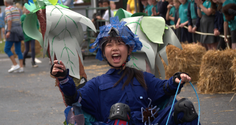 Girl in a customised go-cart style car, cheering. She's dressed in a blue overall and decorated racing helmet. Representing DT projects Year 6