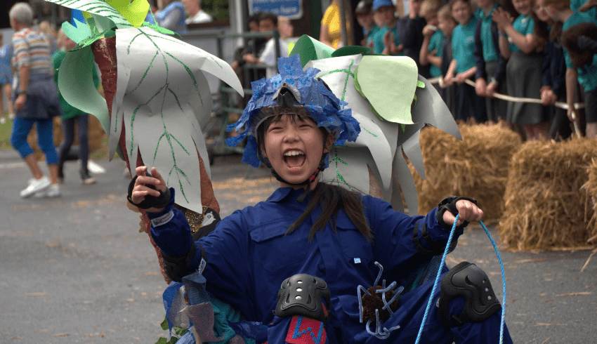 Girl in a customised go-cart style car, cheering. She's dressed in a blue overall and decorated racing helmet. Representing DT projects Year 6