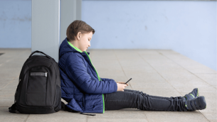Boy with schoolbag sitting on floor looking at phone, representing poor school attendance