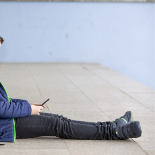 Boy with schoolbag sitting on floor looking at phone, representing poor school attendance