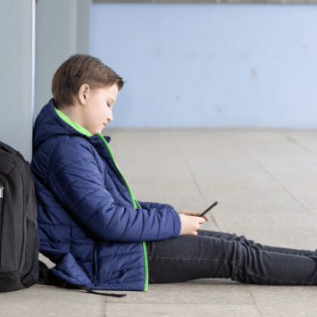 Boy with schoolbag sitting on floor looking at phone, representing poor school attendance