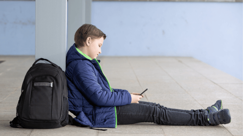 Boy with schoolbag sitting on floor looking at phone, representing poor school attendance