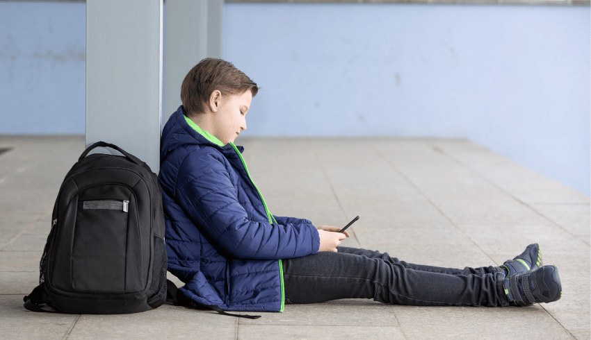 Boy with schoolbag sitting on floor looking at phone, representing poor school attendance