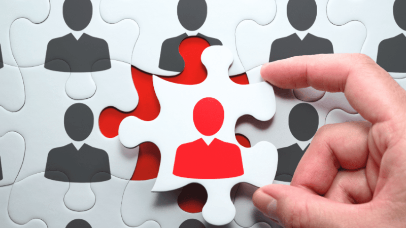 Hand selecting a puzzle piece with a red head and shoulders on it, representing teacher recruitment