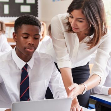 A secondary school boy using a laptop speaks to a female teacher