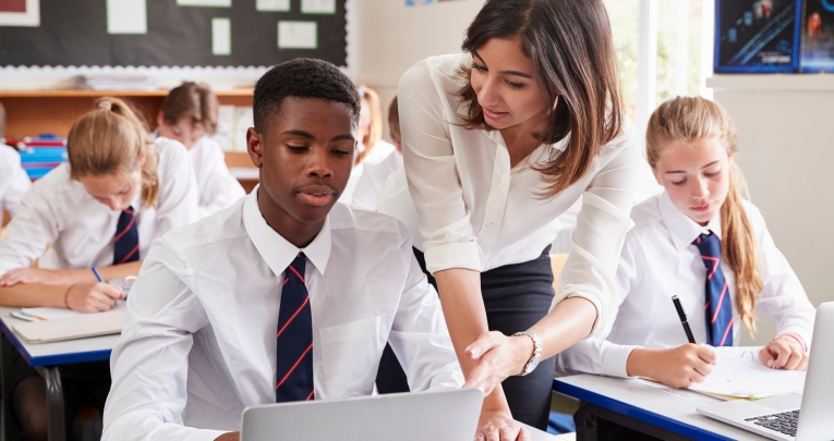 A secondary school boy using a laptop speaks to a female teacher