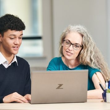 A male secondary school student sitting at a laptop with a tutor