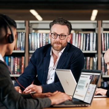 Two secondary school students sitting at a desk in a library with a teacher