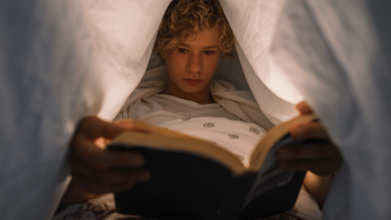 Teenage boy reading book under covers, representing school reading strategy